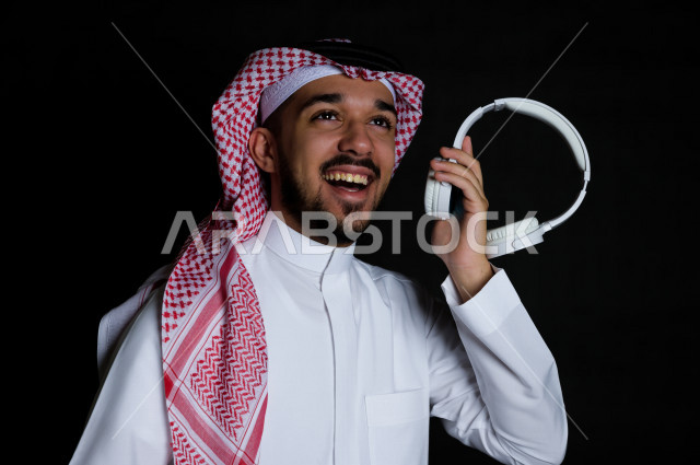 بورتريه لشاب سعودي خليجي عربي يمسك بيده سماعة لاسلكية بيضاء , خلفية سوداء 