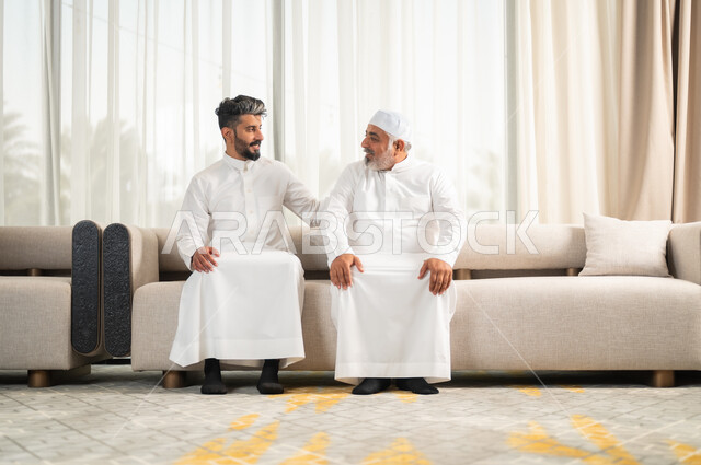 تعابير المودة والمحبة، الاستمتاع بقضاء وقت مع الابناء، رجل كبير بالسن عربي خليجي سعودي يرتدي الطاقية البيضاء والثوب التقليدي يجلس على الاريكة بجانب ابنه الشاب في غرفة الجلوس، التسامر وتبادل الاحاديث، جلسات عائلية سعيدة