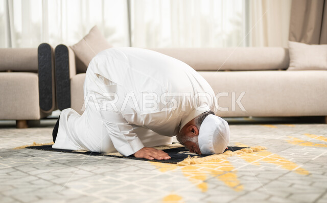 اقامة الصلوات على وقتها والإكثار من الدعاء وذكر الله تعالى، رجل كبير بالسن عربي سعودي خليجي مسلم يسجد على سجادة الصلاة في غرفة المعيشة بخشوع وتضرع، الاجتهاد في العبادة واداء الفرائض الدينية في شهر رمضان المبارك