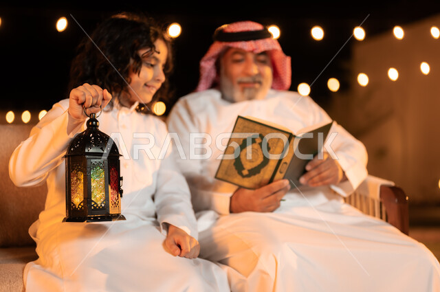 تربية وتعليم الأبناء على العبادة والتقرب من الله تعالى, رجل كبير بالسن عربي خليجي سعودي يرتدي الزي التقليدي يحمل بيده القرآن الكريم ويجلس في حديقة المنزل برفقة حفيده، فوانيس وزينة الاعياد والمناسبات الاسلامية, تلاوة وحفظ المصحف الشريف في شهر رمضان المبارك
