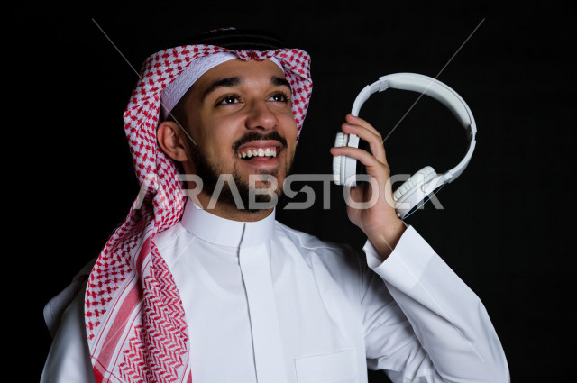 Portrait of a young Saudi Arabian Gulf Arab holding a  White wireless headphones , black background 