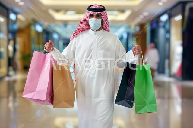 اتخاذ الاجراءات الاحترازية للوقاية من الامراض، متعة التسوق من المولات والمراكز التجارية، رجل عربي خليجي سعودي يرتدي الشماغ والثوب التقليدي يضع الكمامة يحمل بيده أكياس ورقية ملونة، التسوق واقتناء المستلزمات والهدايا، الاستفادة من عروض وتخفيضات الجمعة البيضاء