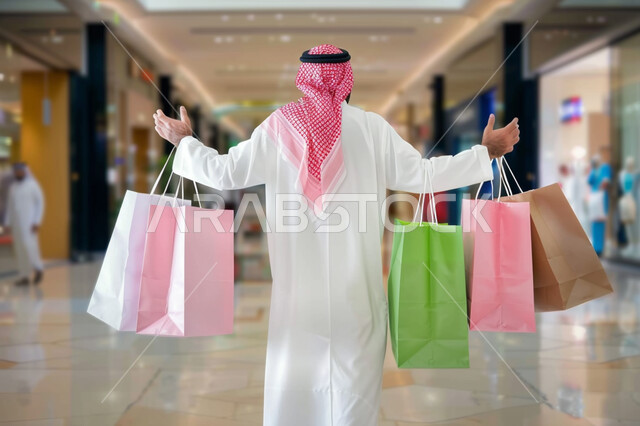 تعابير وحركات الحماس للتسوق واقتناء المستلزمات والهدايا، الاستفادة من عروض وتخفيضات الجمعة البيضاء، شراء الأغراض ومتعة التسوق في المولات والمحلات التجارية، صورة من الخلف لرجل عربي خليجي سعودي يرتدي الشماغ والثوب التقليدي يحمل بيده أكياس ورقية ملونة