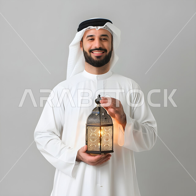 المناسبات الدينية والأعياد الإسلامية, الاستعداد لقدوم شهر رمضان, بورتريه لشاب عربي خليجي سعودي يرتدي الثوب و الغترة التقليدية يحمل الفانوس المضيء وينظر الى الكاميرا بتعابير الفرح, تحف وزينة الاحتفالات في السعودية,  استقبال شهر الخير والبركة, خلفية رمادية