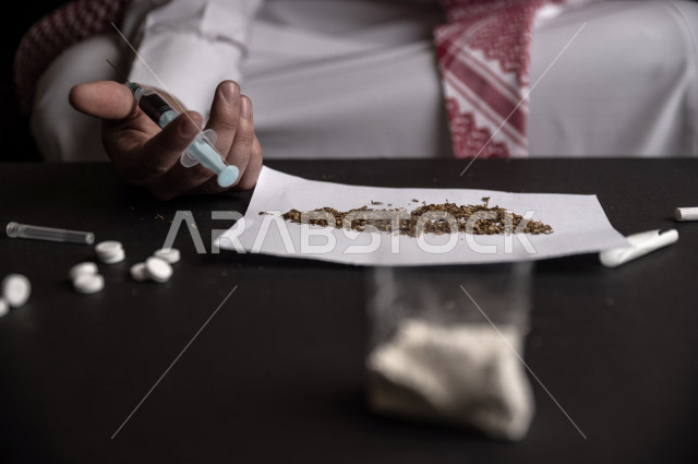 شاب عربي سعودي يعاني من الادمان على المواد الضارة ،  يجلس امام انواع عديدة من المواد السامة  ،  ، الادمان على التبغ ، ادمان السجائر ، تعاطي مواد ضارة و سامة ،  الهيروين والكوكائين، عادات سلوكية ضارة ، لا للتدخين