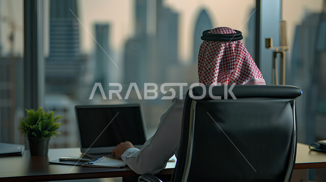 مفهوم إدارة شؤون الأعمال وتنظيمها عبر الاجهزة التقنية، دمج العمل مع التقنية والتكنولوجيا، مهن ووظائف ادارية مكتبية, صورة من الخلف لرجل اعمال عربي خليجي سعودي يقوم بالعمل على الحاسوب المحمول داخل المكتب، واجهة زجاجية مطلة على الأبراج وناطحات السحاب
