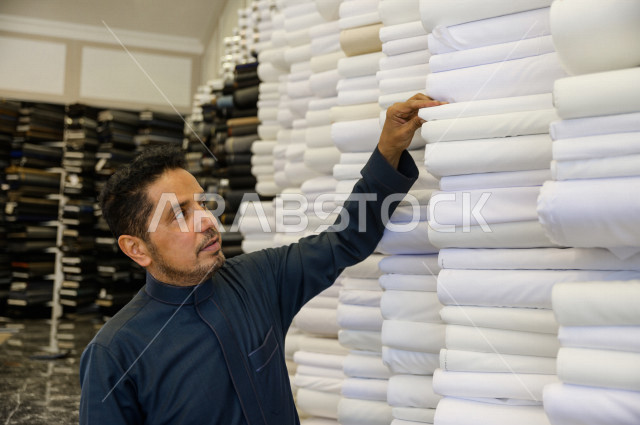 شاب سعودي خليجي يعمل في مهنة الخياطة ، يعرض للزبون اجود انواع الأقنشة لتفصيل الثوب السعودي ، محل خياطة و تفصيل الملابس الرجالي في السعودية ، مهارة الخياطة واتقان تفصيل الملابس الشبابية