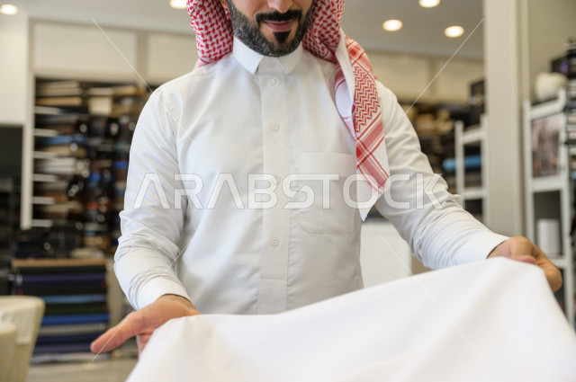 شاب سعودي خليجي داخل محل مختص بخياطة الملابس الرجالية ، اختيار اجود انواع الأقنشة لتفصيل الثوب السعودي ، محل خياطة و تفصيل الملابس الرجالي في السعودية ، مهارة الخياطة واتقان تفصيل الملابس الشبابية