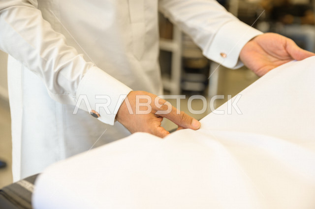 شاب سعودي خليجي داخل محل مختص بخياطة الملابس الرجالية ، اختيار اجود انواع الأقنشة لتفصيل الثوب السعودي ، محل خياطة و تفصيل الملابس الرجالي في السعودية ، مهارة الخياطة واتقان تفصيل الملابس الشبابية