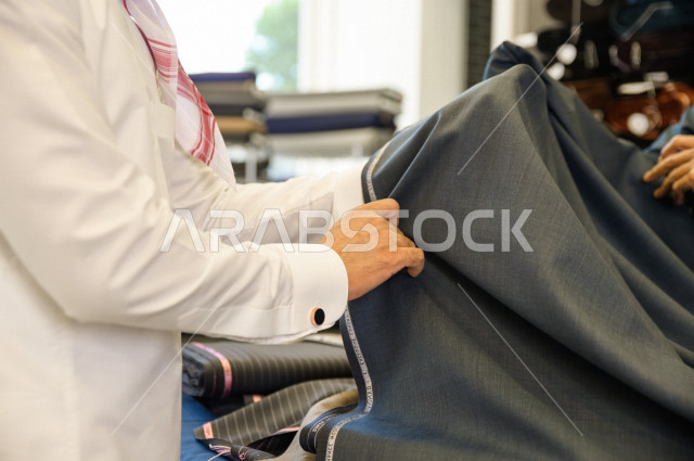 A young Saudi Gulf man inside a shop specializing in sewing men's clothes, choosing the finest types of fabrics for tailoring the Saudi dress, sewing and tailoring men's clothes in Saudi Arabia, sewing skill and mastering the tailoring of youth clothes