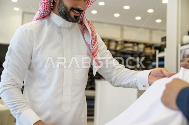 شاب سعودي خليجي يعمل في مهنة الخياطة ، يعرض للزبون اجود انواع الأقنشة لتفصيل الثوب السعودي ، محل خياطة و تفصيل الملابس الرجالي في السعودية ، مهارة الخياطة واتقان تفصيل الملابس الشبابية