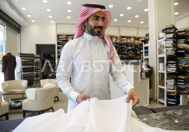شاب سعودي خليجي داخل محل مختص بخياطة الملابس الرجالية ، اختيار اجود انواع الأقنشة لتفصيل الثوب السعودي ، محل خياطة و تفصيل الملابس الرجالي في السعودية ، مهارة الخياطة واتقان تفصيل الملابس الشبابية