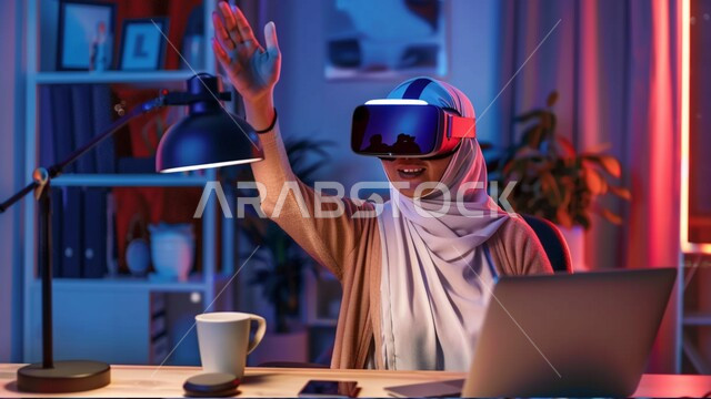 Expressions and gestures of integration and focus in virtual reality, simulating the imaginary world, using advanced modern devices and techniques, a Saudi Gulf Arab girl wearing the hijab and VR glasses sitting at her desk in front of the laptop, raising her hand upwards, digital transformation and technical progress.