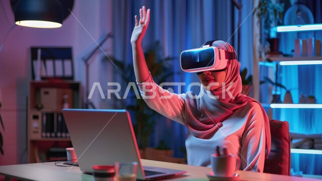 Using advanced modern devices and techniques, a Saudi Arabian Gulf girl wearing a hijab and VR glasses sitting at her desk in front of the laptop, raising her hand upwards, digital transformation and technical progress, expressions and gestures of integration and focus in virtual reality, simulating the imaginary world.