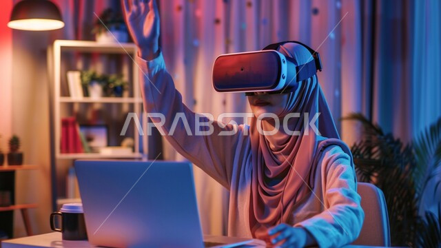 التحول الرقمي والتقدم التقني، تعابير وايماءات الاندماج والتركيز في الواقع الافتراضي، محاكاة العالم الخيالي، استخدام الأجهزة والتفنيات الحديثة المتطورة، فتاة عربية خليجية سعودية ترتدي الحجاب ونظارات الفي آر تجلس على مكتبها امام الحاسوب المحمول ترفع يدها الى الاعلى