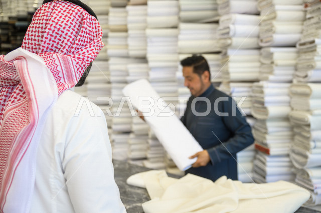 شاب سعودي خليجي يعمل في مهنة الخياطة ، يعرض للزبون اجود انواع الأقنشة لتفصيل الثوب السعودي ، محل خياطة و تفصيل الملابس الرجالي في السعودية ، مهارة الخياطة واتقان تفصيل الملابس الشبابية