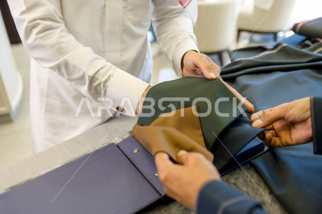 شاب سعودي خليجي يعمل في مهنة الخياطة ، يعرض للزبون اجود انواع الأقنشة لتفصيل الثوب السعودي ، محل خياطة و تفصيل الملابس الرجالي في السعودية ، مهارة الخياطة واتقان تفصيل الملابس الشبابية