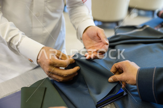 شاب سعودي خليجي يعمل في مهنة الخياطة ، يعرض للزبون اجود انواع الأقنشة لتفصيل الثوب السعودي ، محل خياطة و تفصيل الملابس الرجالي في السعودية ، مهارة الخياطة واتقان تفصيل الملابس الشبابية