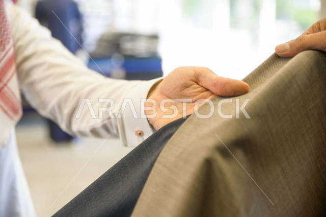 A young Saudi Gulf man working in the profession of sewing, offering to the customer the finest types of fabrics for tailoring the Saudi dress, a shop for sewing and tailoring men's clothes in Saudi Arabia, the skill of sewing and mastering the tailoring 