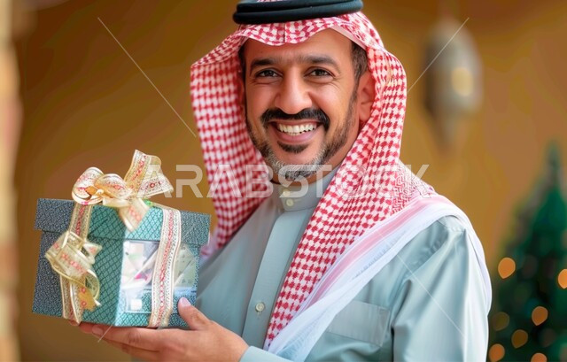 أعياد ومناسبات ومفاجآت سعيدة, إهداء وتقديم الهدايا الفاخرة، صورة مقربة لرجل عربي خليجي سعودي مبتسم يرتدي الثوب والشماغ التقليدي يحمل بيديه علبة هدايا مغلفة ينظر الى الكاميرا بإيماءات السرور والسعادة