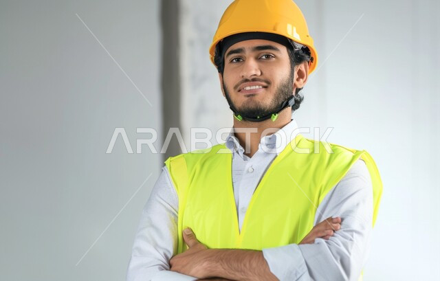 الإعمار بسواعد أبناء الوطن، مهن ووظائف سعودية، مفهوم الهندسة والانشاءات المعمارية، صورة مقربة لمهندس عربي خليجي سعودي مبتسم يرتدي الخوذه  وسترة الحماية ينظر إلى شيء ما بإيماءات السرور، الوقوف بأيدي متقاطعه، العمل في القطاع الهندسي
