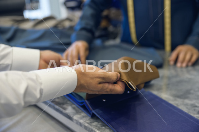 شاب سعودي خليجي يعمل في مهنة الخياطة ، يعرض للزبون اجود انواع الأقنشة لتفصيل الثوب السعودي ، محل خياطة و تفصيل الملابس الرجالي في السعودية ، مهارة الخياطة واتقان تفصيل الملابس الشبابية