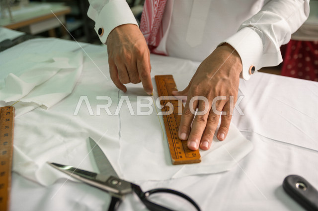 شاب سعودي خليجي يعمل في مهنة الخياطة ، يستخدم الأدوات المناسبة لتفصيل جلابية خاصة لاحدى الزبائن ، محل خياطة و تفصيل الملابس الرجالي في السعودية ، مهارة الخياطة واتقان تفصيل الملابس الشبابية