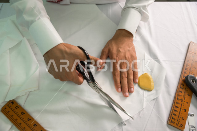 شاب سعودي خليجي يعمل في مهنة الخياطة ، يستخدم الأدوات المناسبة لتفصيل جلابية خاصة لاحدى الزبائن ، محل خياطة و تفصيل الملابس الرجالي في السعودية ، مهارة الخياطة واتقان تفصيل الملابس الشبابية