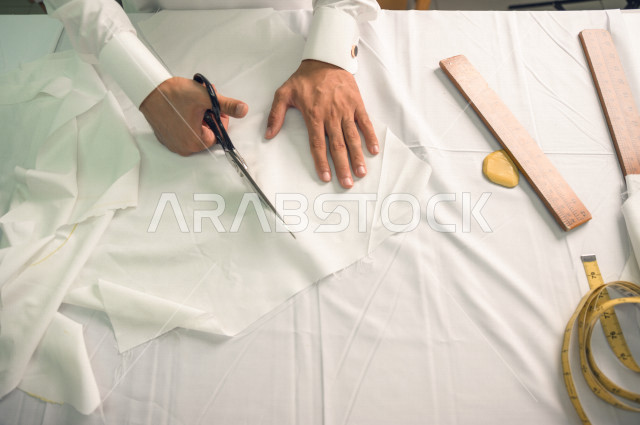 شاب سعودي خليجي يعمل في مهنة الخياطة ، يستخدم الأدوات المناسبة لتفصيل جلابية خاصة لاحدى الزبائن ، محل خياطة و تفصيل الملابس الرجالي في السعودية ، مهارة الخياطة واتقان تفصيل الملابس الشبابية