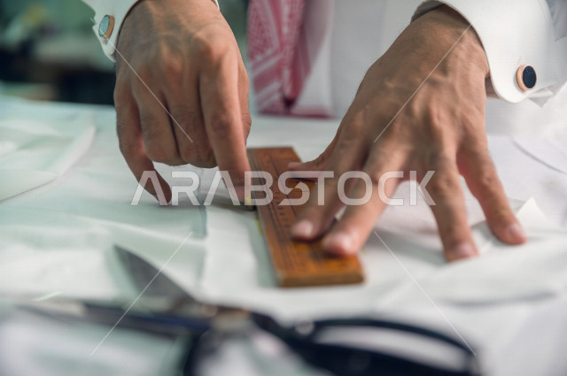 شاب سعودي خليجي يعمل في مهنة الخياطة ، يستخدم الأدوات المناسبة لتفصيل جلابية خاصة لاحدى الزبائن ، محل خياطة و تفصيل الملابس الرجالي في السعودية ، مهارة الخياطة واتقان تفصيل الملابس الشبابية