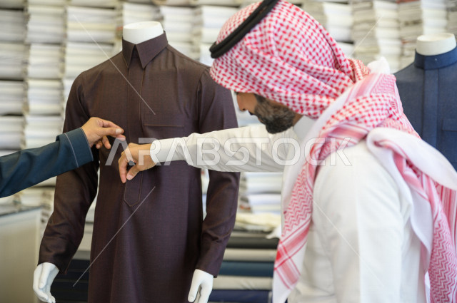 شاب سعودي خليجي يعمل في مهنة الخياطة ، يقوم بأخذ القياسات الخاصة بجسم الزبون من حيث الطول و العرض و الخصر باستخدام الشريط المتري ، محل خياطة و تفصيل الملابس الرجالي في السعودية ، مهارة الخياطة واتقان تفصيل الملابس الشبابية