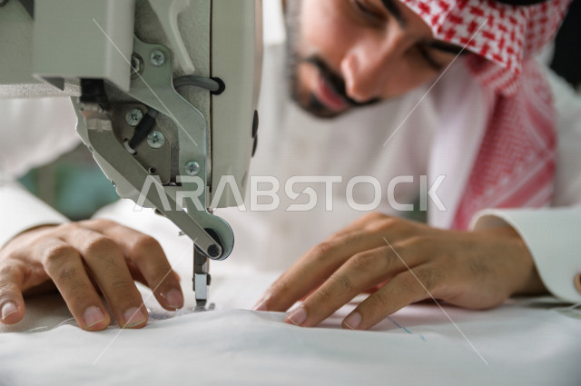 شاب سعودي خليجي يعمل في مهنة الخياطة ، يستخدم الأدوات المناسبة لتفصيل جلابية خاصة لاحدى الزبائن ، محل خياطة و تفصيل الملابس الرجالي في السعودية ، مهارة الخياطة واتقان تفصيل الملابس الشبابية