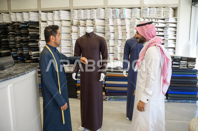 شاب سعودي خليجي يعمل في مهنة الخياطة ، يقوم بأخذ القياسات الخاصة بجسم الزبون من حيث الطول و العرض و الخصر باستخدام الشريط المتري ، محل خياطة و تفصيل الملابس الرجالي في السعودية ، مهارة الخياطة واتقان تفصيل الملابس الشبابية