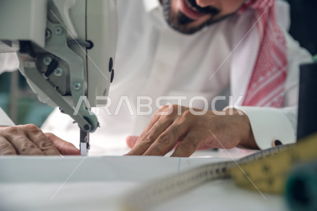 شاب سعودي خليجي يعمل في مهنة الخياطة ، يستخدم الأدوات المناسبة لتفصيل جلابية خاصة لاحدى الزبائن ، محل خياطة و تفصيل الملابس الرجالي في السعودية ، مهارة الخياطة واتقان تفصيل الملابس الشبابية