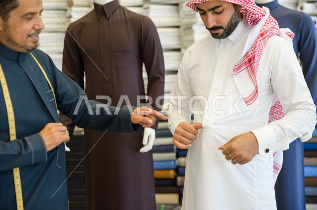 شاب سعودي خليجي يعمل في مهنة الخياطة ، يقوم بأخذ القياسات الخاصة بجسم الزبون من حيث الطول و العرض و الخصر باستخدام الشريط المتري ، محل خياطة و تفصيل الملابس الرجالي في السعودية ، مهارة الخياطة واتقان تفصيل الملابس الشبابية