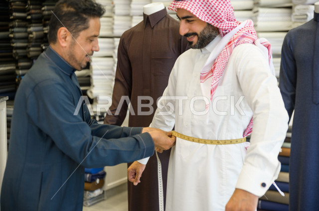 شاب سعودي خليجي يعمل في مهنة الخياطة ، يقوم بأخذ القياسات الخاصة بجسم الزبون من حيث الطول و العرض و الخصر باستخدام الشريط المتري ، محل خياطة و تفصيل الملابس الرجالي في السعودية ، مهارة الخياطة واتقان تفصيل الملابس الشبابية