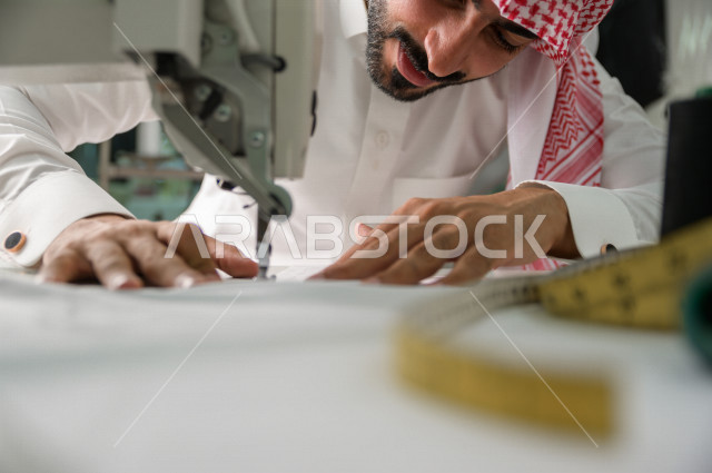 شاب سعودي خليجي يعمل في مهنة الخياطة ، يستخدم الأدوات المناسبة لتفصيل جلابية خاصة لاحدى الزبائن ، محل خياطة و تفصيل الملابس الرجالي في السعودية ، مهارة الخياطة واتقان تفصيل الملابس الشبابية