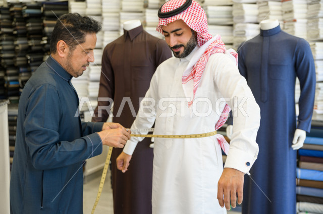 شاب سعودي خليجي يعمل في مهنة الخياطة ، يقوم بأخذ القياسات الخاصة بجسم الزبون من حيث الطول و العرض و الخصر باستخدام الشريط المتري ، محل خياطة و تفصيل الملابس الرجالي في السعودية ، مهارة الخياطة واتقان تفصيل الملابس الشبابية