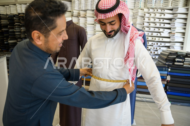 A young Saudi Gulf man working in the profession of sewing, he takes measurements of the customer's body in terms of length, width and waist using metric tape, a shop for sewing and tailoring men's clothes in Saudi Arabia, sewing skill and mastering the d