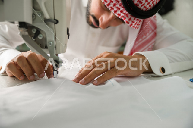 شاب سعودي خليجي يعمل في مهنة الخياطة ، يستخدم الأدوات المناسبة لتفصيل جلابية خاصة لاحدى الزبائن ، محل خياطة و تفصيل الملابس الرجالي في السعودية ، مهارة الخياطة واتقان تفصيل الملابس الشبابية