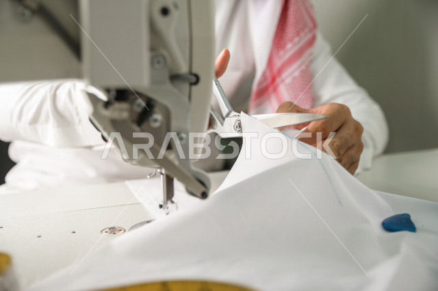 شاب سعودي خليجي يعمل في مهنة الخياطة ، يستخدم الأدوات المناسبة لتفصيل جلابية خاصة لاحدى الزبائن ، محل خياطة و تفصيل الملابس الرجالي في السعودية ، مهارة الخياطة واتقان تفصيل الملابس الشبابية