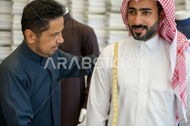 شاب سعودي خليجي يعمل في مهنة الخياطة ، يقوم بأخذ القياسات الخاصة بجسم الزبون من حيث الطول و العرض و الخصر باستخدام الشريط المتري ، محل خياطة و تفصيل الملابس الرجالي في السعودية ، مهارة الخياطة واتقان تفصيل الملابس الشبابية