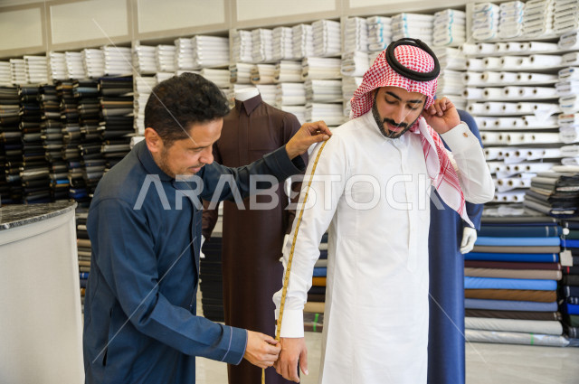 شاب سعودي خليجي يعمل في مهنة الخياطة ، يقوم بأخذ القياسات الخاصة بجسم الزبون من حيث الطول و العرض و الخصر باستخدام الشريط المتري ، محل خياطة و تفصيل الملابس الرجالي في السعودية ، مهارة الخياطة واتقان تفصيل الملابس الشبابية