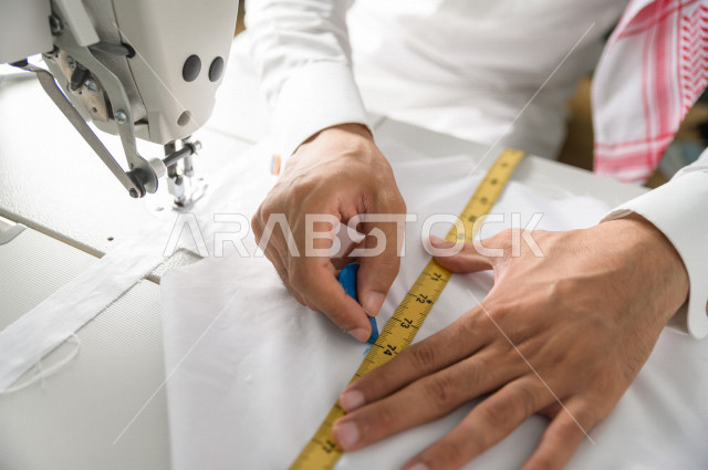 شاب سعودي خليجي يعمل في مهنة الخياطة ، يستخدم الأدوات المناسبة لتفصيل جلابية خاصة لاحدى الزبائن ، محل خياطة و تفصيل الملابس الرجالي في السعودية ، مهارة الخياطة واتقان تفصيل الملابس الشبابية