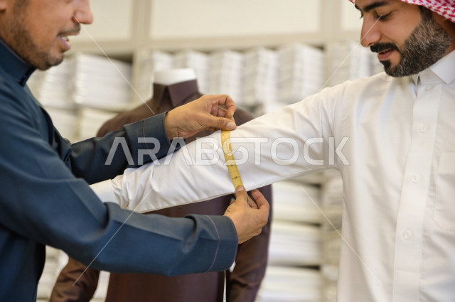 شاب سعودي خليجي يعمل في مهنة الخياطة ، يقوم بأخذ القياسات الخاصة بجسم الزبون من حيث الطول و العرض و الخصر باستخدام الشريط المتري ، محل خياطة و تفصيل الملابس الرجالي في السعودية ، مهارة الخياطة واتقان تفصيل الملابس الشبابية