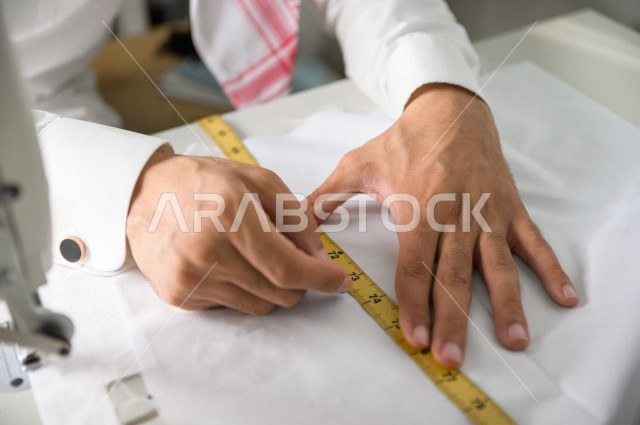 شاب سعودي خليجي يعمل في مهنة الخياطة ، يستخدم الأدوات المناسبة لتفصيل جلابية خاصة لاحدى الزبائن ، محل خياطة و تفصيل الملابس الرجالي في السعودية ، مهارة الخياطة واتقان تفصيل الملابس الشبابية