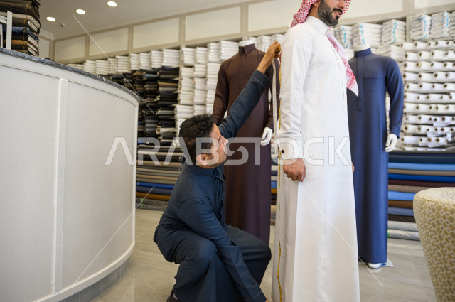 شاب سعودي خليجي يعمل في مهنة الخياطة ، يقوم بأخذ القياسات الخاصة بجسم الزبون من حيث الطول و العرض و الخصر باستخدام الشريط المتري ، محل خياطة و تفصيل الملابس الرجالي في السعودية ، مهارة الخياطة واتقان تفصيل الملابس الشبابية