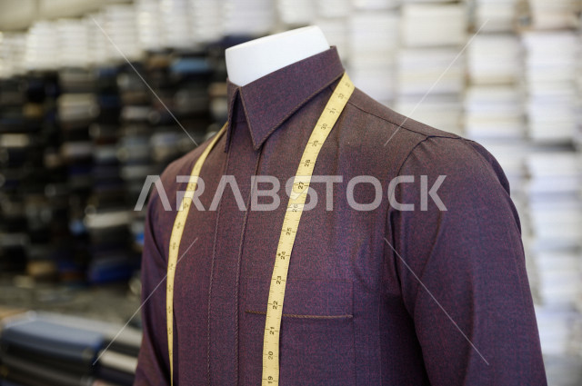 شاب سعودي خليجي يعمل في مهنة الخياطة ، يقوم بأخذ القياسات الخاصة بجسم الزبون من حيث الطول و العرض و الخصر باستخدام الشريط المتري ، محل خياطة و تفصيل الملابس الرجالي في السعودية ، مهارة الخياطة واتقان تفصيل الملابس الشبابية