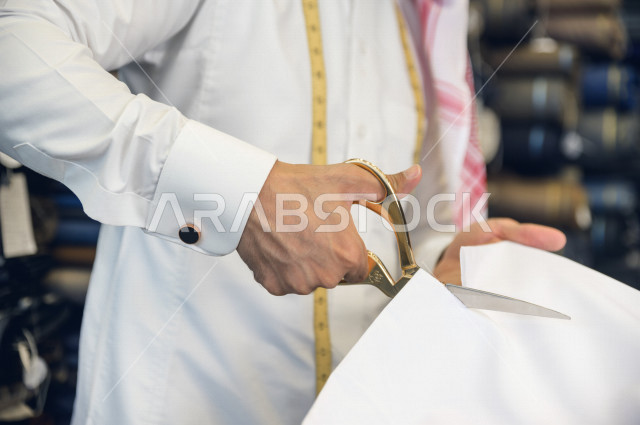 شاب سعودي خليجي يعمل في مهنة الخياطة ، يستخدم الأدوات المناسبة لتفصيل جلابية خاصة لاحدى الزبائن ، محل خياطة و تفصيل الملابس الرجالي في السعودية ، مهارة الخياطة واتقان تفصيل الملابس الشبابية