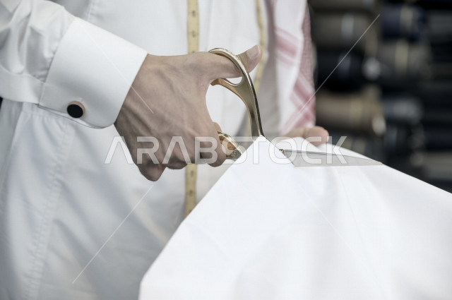 شاب سعودي خليجي يعمل في مهنة الخياطة ، يستخدم الأدوات المناسبة لتفصيل جلابية خاصة لاحدى الزبائن ، محل خياطة و تفصيل الملابس الرجالي في السعودية ، مهارة الخياطة واتقان تفصيل الملابس الشبابية